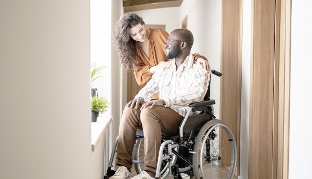 Junger Mann im Rollstuhl vor Fenster im Hausflur mit Partnerin
