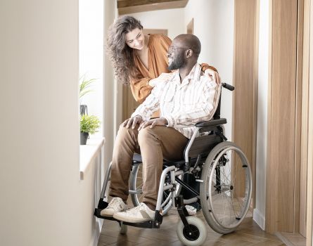 Junger Mann im Rollstuhl vor Fenster im Hausflur mit Partnerin